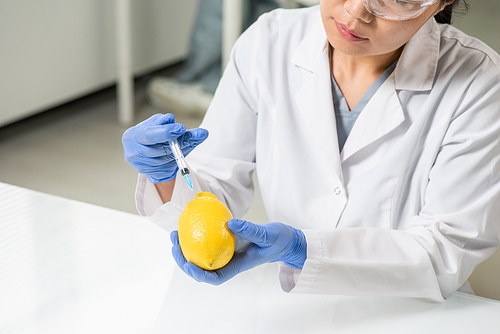Gloved hands of contemporary young female scientist in whitecoat injecting lemon over table while making experiment in laboratory
