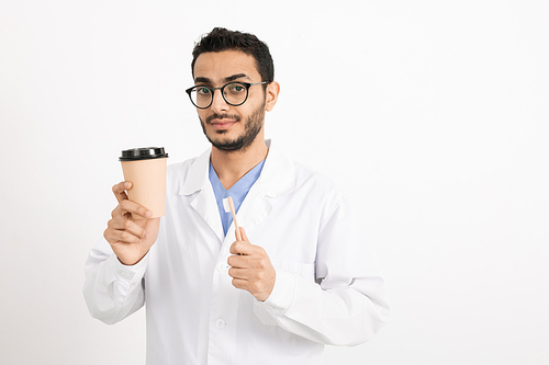 Young male dentist in whitecoat and eyeglasses holding glass of coffee and toothbrush while looking at you in isolation