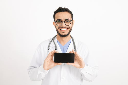 Cheerful young doctor with toothy smile photographing you on smartphone in front of camera in isolation