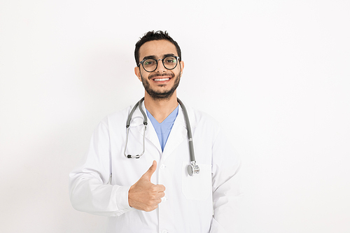 Young smiling clinician in eyeglasses and whitecoat showing thumb up while standing in front of camera in isolation