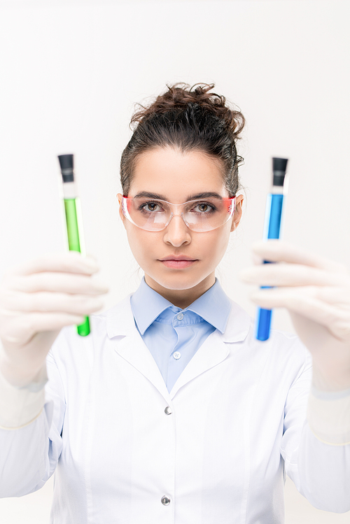 Contemporary female clinician or chemist in gloves and protective eyewear showing flasks with blue and green fluids