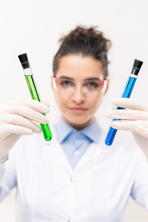 Gloved hands of female clinician or pharmacist showing flasks with blue and green vaccine samples in laboratory