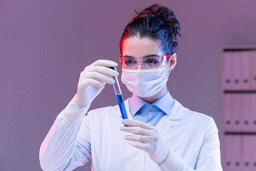 Young female scientist in mask, gloves and eyeglasses looking at flask while studying blue liquid substance in laboratory