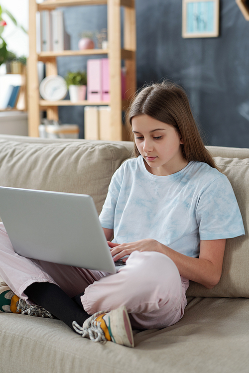 Serious teenage girl in homewear sitting with crossed legs on sofa and using laptop while communicating online