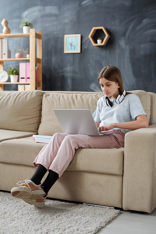 Cute female teenage student in casualwear looking at laptop display while relaxing on couch in living-room after school