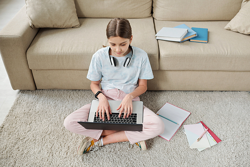 Above view of concentrated teenage girl sitting on carpet and using laptop while learning lesson