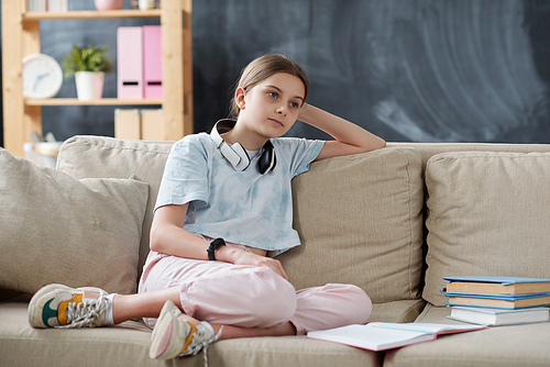 Pensive attractive teenage girl with wireless headphones around neck sitting on sofa with textbooks