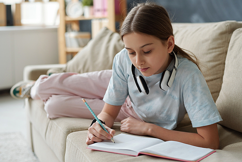 Concentrated pretty girl with wireless headphones on neck lying on sofa and solving school task in workbook