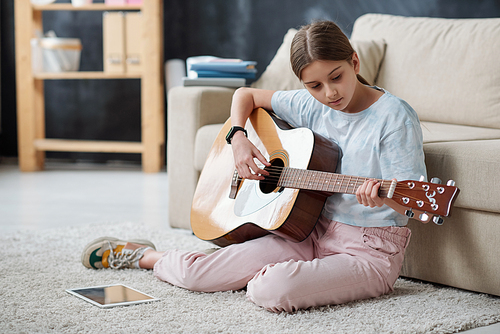 Self-taught teenage girl sitting on carpet and using instructional video on tablet while playing guitar