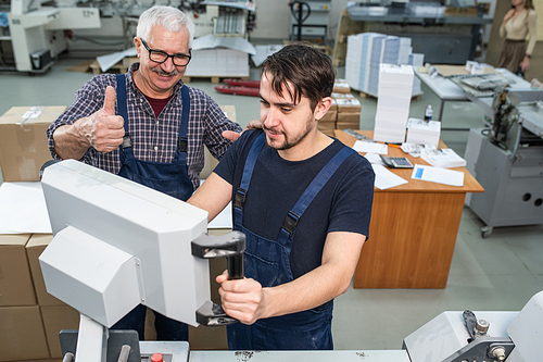 Satisfied senior man showing thumb-up while giving assessment to young intern operating machine at printing plant