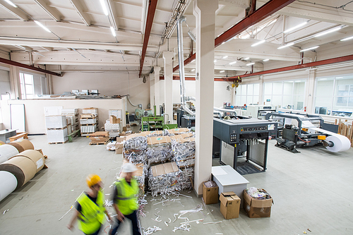 Interior of printing house with packaged paper garbage, printing machines and paper rolls, blurred motion of workers