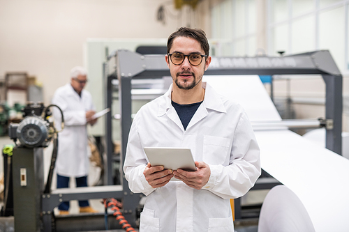 Portrait of young printing house engineer in white coat and glasses holding digital tablet in factory shop