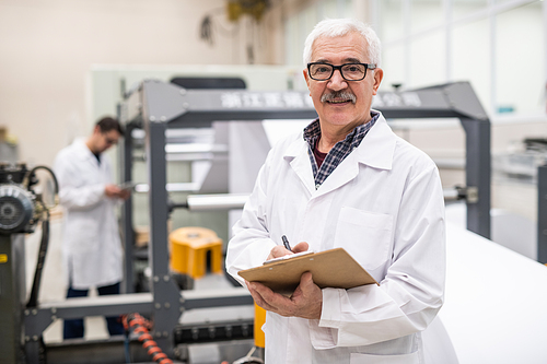 Portrait of smiling senior quality inspector in lab coat making notes in clipboard at factory