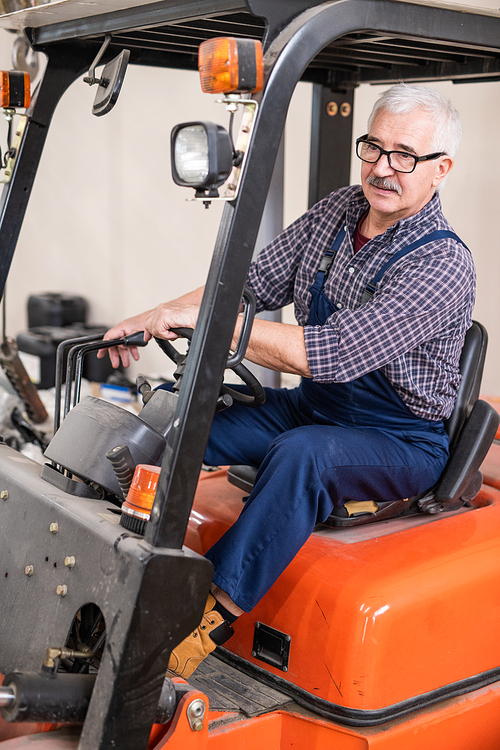 Serious senior forklift truck driver in glasses loading goods from deliveries at factory