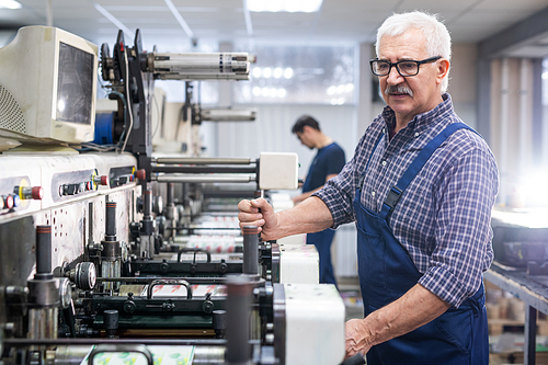 Serious senior white-haired man in overall operating printing press machine at factory