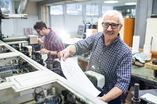 Portrait of cheerful senior man in glasses getting printed paper out of printing press at factory