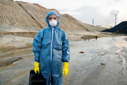 Young female scientist in protective coveralls, rubber gloves and respirator holding briefcase with water and soil samples standing against hills