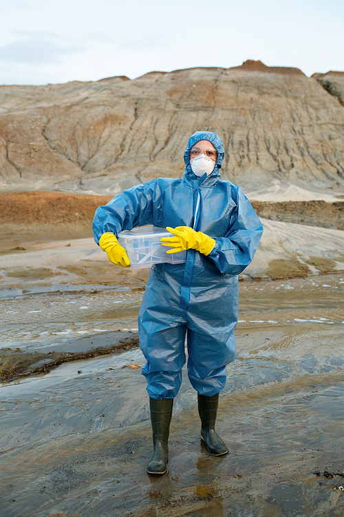 Young female scientist in protective coveralls, respirator, rubber gloves and boots holding plastic container with water and soil samples