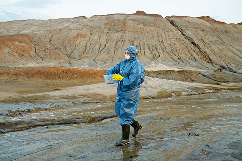 Contemporary female scientist in protective coveralls, respirator, rubber gloves and boots carrying plastic container against hills and sky