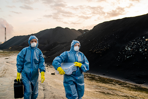 Two female ecologists in coveralls, respirators, eyeglasses and rubber gloves carrying cases with soil samples while moving along road