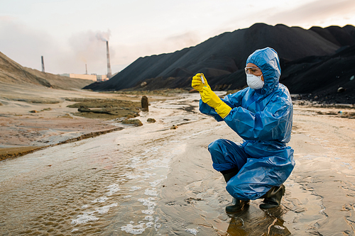 Contemporary young female ecologist or researcher in protective workwear and respirator putting sample of soil into flask in toxic area