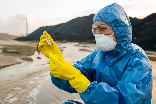 Young female ecologist or researcher in protective coveralls, respirator and eyeglasses looking at flask with sample of soil in toxic area