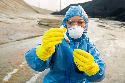 Young female ecologist in respirator and eyeglasses looking at flask with sample of dirty water in polluted and toxic industrial area