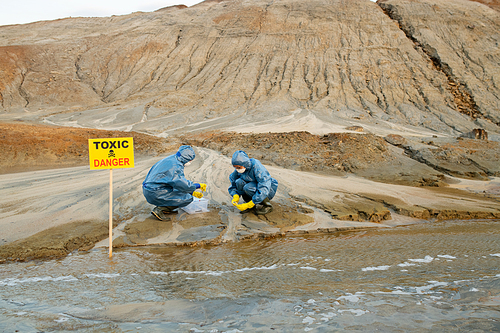Ecologists in rubber gloves and boots, protective coveralls and respirators taking samples of toxic soil and water in polluted area against hills
