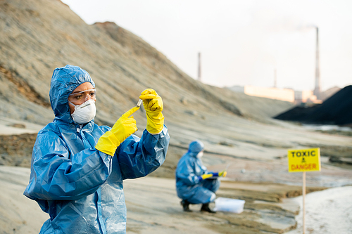 Young researcher in protective coveralls looking at sample of toxic soil in her hands while making investigation with colleague in dangerous area
