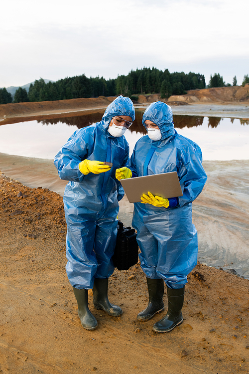 Young female ecologists in protective workwear standing on polluted soil or clay against dirty river and making notes in laptop