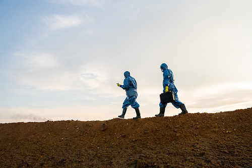 Two contemporary researchers in protective coveralls moving on top of hill with dirty soil against cloudy sky while carrying out investigation