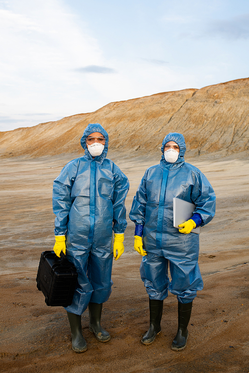 Two young female researchers in protective workwear standing on polluted soil in abandoned territory against hill and sky during investigation