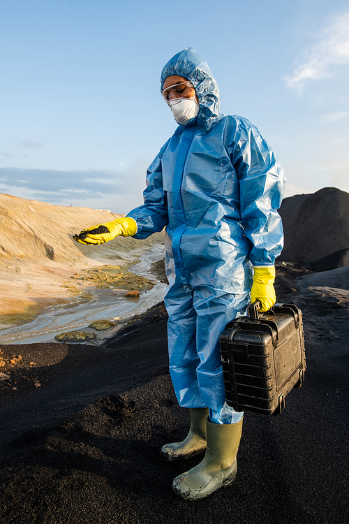 Young female ecologist in protective workwear holding briefcase with samples of polluted water and soil while standing on top of hill