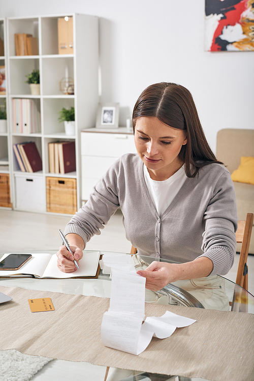 Serious attractive young woman sitting at table and viewing financial check while recording expenses in diary