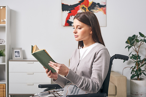 Young disable brunette woman in casualwear sitting in wheelchair and reading book while spending day at home