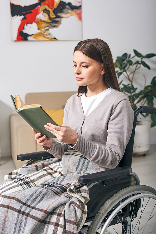 Attractive young disabled woman sitting under plaid in wheelchair and reading book at home