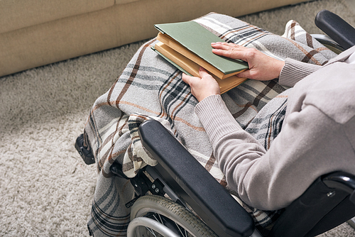 Hands of young disable woman holding book on her knees while sitting in wheelchair and spending time at home