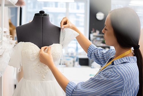 Serious female tailor with ponytail putting wedding dress on dummy and thinking of design of shoulder cover