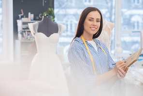 Horizontal medium portrait of young woman standing in wedding dress-making studio holding sketchbook  smiling