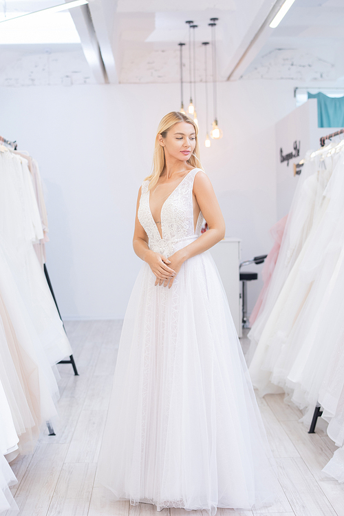 Content elegant young blond-haired bride fitting wedding gown in shop