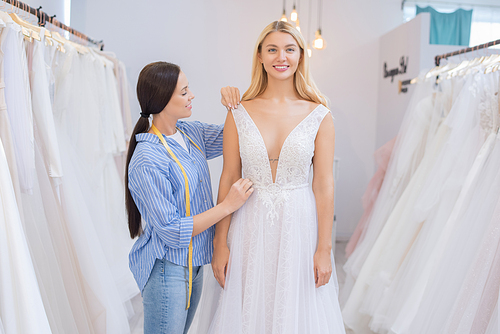 Smiling wedding dress tailor with tape measure adjusting gown fitted by young bride in wedding show room