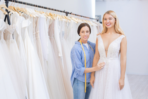 Attractive young woman trying on wedding dress in modern bridal shop, sales assistant helping her