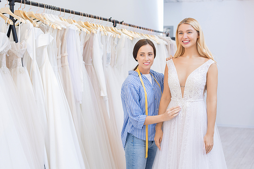 Attractive young woman trying on wedding dress in modern bridal shop, sales assistant helping her