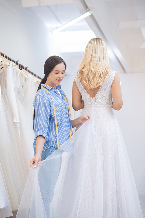 Content young dressmaker adjusting hem of wedding dress during gown fitting in studio