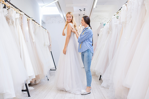 Female shop assistant helping young woman to fasten wedding dress in modern bridal store, horizontal shot
