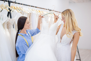 Positive blond-haired bride choosing wedding dress for fitting with consultant in shop