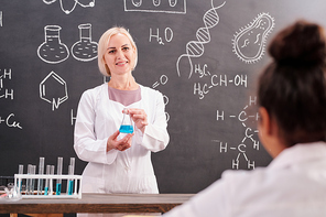 Young blond smiling teacher of chemistry holding tube with blue liquid substance while standing by blackboard with chemical formula