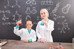 Clever schoolgirl and successful teacher of chemistry in whitecoats holding tubes with red and blue fluids over table while showing reaction