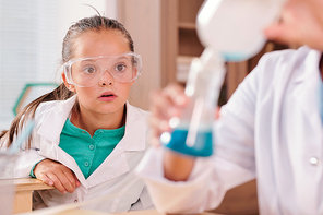 Cute astonished schoolgirl in protective eyeglasses and whitecoat looking at blue liquid substance in large beak held by her classmate at lesson