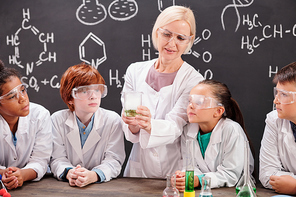 Young blond teacher of chemistry in whitecoat showing her pupils mixture of two chemical substances while discussing reaction together